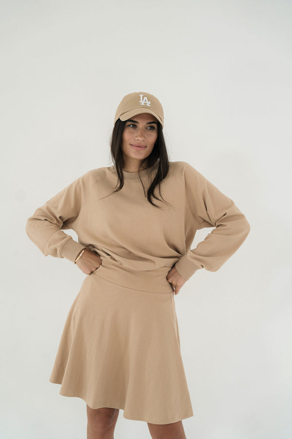 Woman wearing a beige skirt, crewneck top, and cap with a logo on a white background